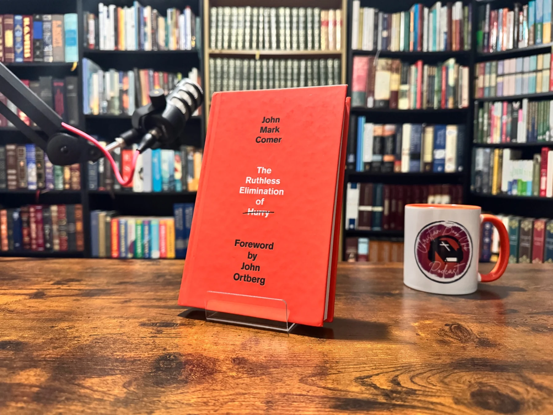 The Ruthless Elimination of Hurry book on a wooden table beside a podcast microphone and branded mug